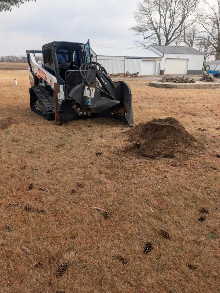 Bobcat compact track loader with a stump grinding attachment working in a grassy yard beside a farm outbuilding